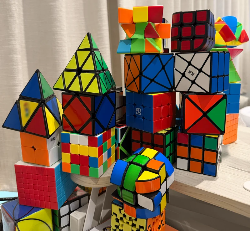 Photograph of about 20 puzzles stacked in 6 towers on a desk, in front of a cream curtain.
